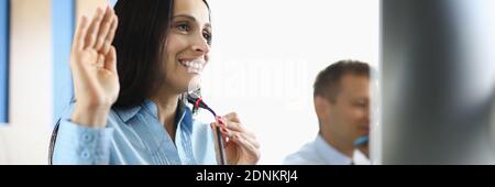 Donna d'affari si siede al computer con il microfono sorridente e sollevando la mano in modo accogliente. Foto Stock