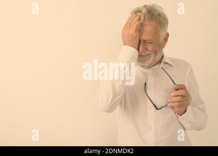 Studio shot di sad senior uomo barbuto tergi le lacrime mentre si tiene un occhiale contro uno sfondo bianco Foto Stock