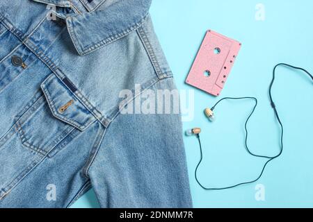 Giacca jeans, audiocassetta, auricolari sottovuoto su sfondo blu pastello. Retrò media, amante della musica, anni '80. Vista dall'alto, disposizione piatta Foto Stock