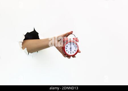 Una mano femminile con manicure rossa attraverso la carta bianca strappata tiene una sveglia rossa. Passare la mano attraverso la carta bianca strappata. Mano che tiene una sveglia rossa Foto Stock