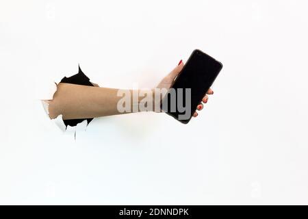 Una mano femminile con una manicure rossa attraverso la carta bianca strappata tiene uno smartphone mobile con uno schermo nero vuoto. Passare la mano attraverso la carta bianca strappata Foto Stock