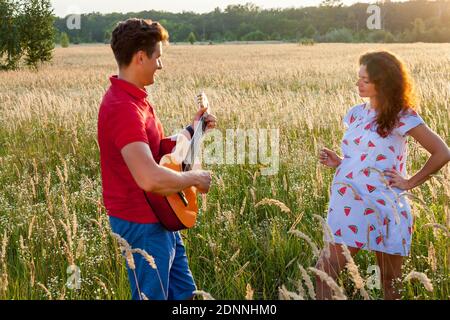 Un giovane con la chitarra è in piedi vicino alla moglie incinta nel campo di grano in una giornata di sole. Foto Stock