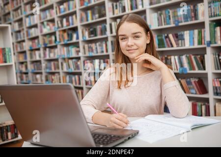 Allegra ragazza adolescente sorridente alla macchina fotografica mentre lavora sul suo laptop alla biblioteca dell'università, spazio di copia. E-learning, concetto di istruzione Foto Stock