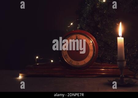 Antico orologio in legno d'epoca, candela gialla e albero di Natale ramo su sfondo. Mezzanotte in orologio. Candela che brucia. Foto Stock