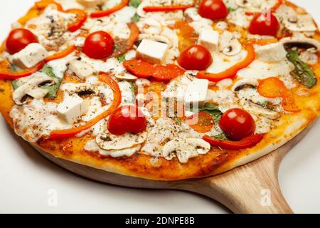 Deliziosa pizza vegetariana servita su un piatto di legno, gli ingredienti sono salsa di firma, basilico, mozzarella, champignons, peperoni, pomodori ciliegini, formaggio feta. Poster promozionale pizzeria. Foto Stock