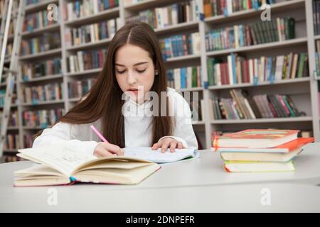 Giovane ragazza teenage che guarda concentrata, scrivendo nel suo libro di testo alla biblioteca, femmina studente teen che lavora per l'assegnazione per la scuola Foto Stock