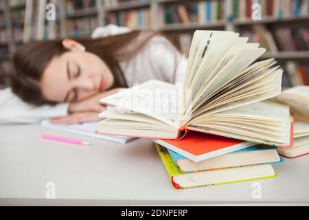 Fuoco selettivo sul mucchio dei libri alla biblioteca, la ragazza teenage che dorme sui suoi fogli di lavoro dopo lo studio alla biblioteca Foto Stock