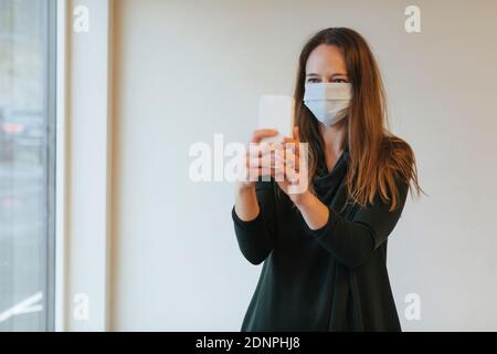 Donna che indossa una maschera protettiva mentre prende selfie Foto Stock