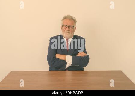 Studio shot di bello senior barbuto imprenditore con le braccia incrociate mentre è seduto su un tavolo di legno contro uno sfondo bianco Foto Stock