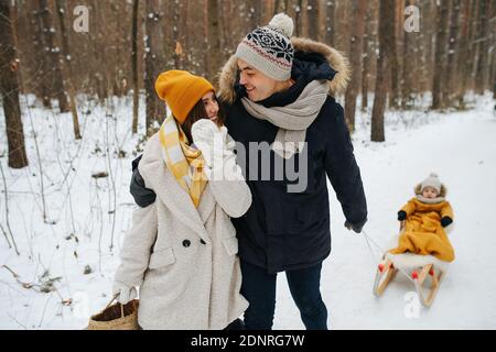 Felice carina famiglia passeggiando in una foresta invernale, papà tirando slitta con un bambino Foto Stock