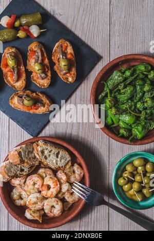 vista dall'alto verticale di un tavolo in legno preparato con casseruola con gamberi e peperoni verdi, un vassoio con piccoli toast con pepe rosso e acciughe e. Foto Stock