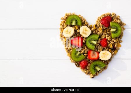 Cuore disteso di granola e frutta su sfondo bianco di legno. Disposizione piatta. Spazio di copia. Foto Stock