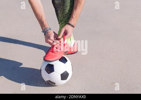 dettaglio di un calciatore che lega le sue scarpe con un piede sulla palla in un campo di calcio in cemento, concetto di stile di vita sano e sport urbano nel ci Foto Stock