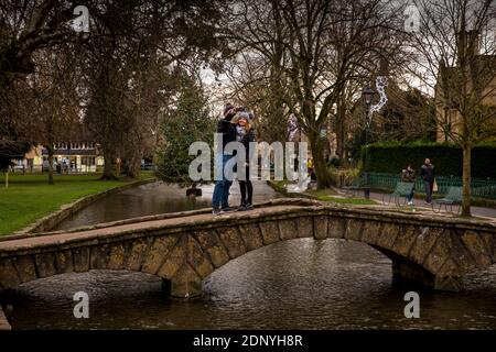 Regno Unito, Gloucestershire, Bourton on the Water, coppia sul ponte prendendo selfie con l'albero di Natale nel fiume Windrush Foto Stock