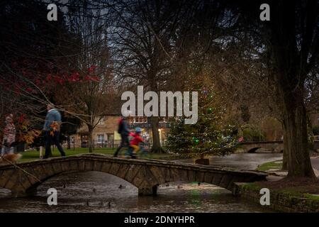 Regno Unito, Gloucestershire, Bourton sull'acqua, persone che attraversano il ponte sul fiume Windrush vicino all'albero di Natale al crepuscolo Foto Stock