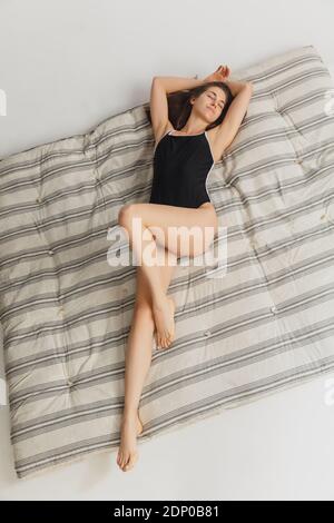 Chill. Beautiful young woman's portrait on grey mattress in studio. Having fun, happy, full length. Dancing, getting crazy happy, having fun. Stylish girl in black sportive swimsuit. Top view. Stock Photo