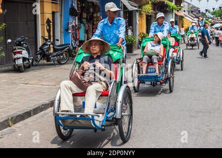 Ha Gai, Vietnam -- 23 marzo 2016. Una linea di turisti in pedicabs cavalcano attraverso una trafficata strada dello shopping ad Hongai, Vietnam. Foto Stock