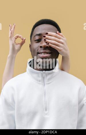 La mano della donna che copre l'occhio dell'uomo sorridente, mostrando il segno ok sullo sfondo Foto Stock