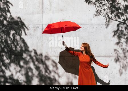 Smiling redhead woman holding umbrella while standing against tree shadow wall Stock Photo