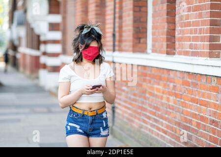 Il viso di una giovane donna è coperto di maschera utilizzando lo smartphone mentre in città Foto Stock