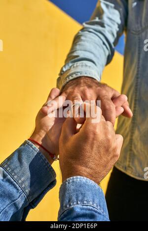 Gay man putting ring in young man finger while standing by yellow wall Foto Stock