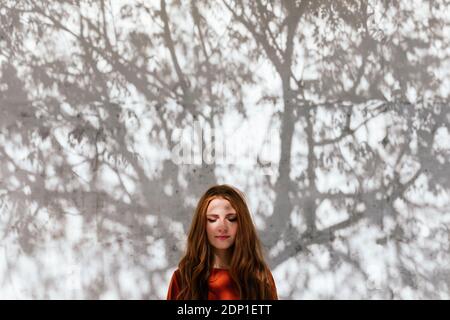 Redhead woman standing with eyes closed against tree shadow wall Stock Photo