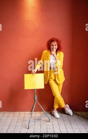 Giovane donna in tuta gialla appoggiata al supporto musicale con dito sulla bocca Foto Stock