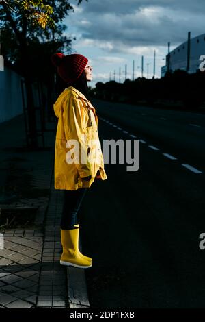 Donna che indossa un impermeabile giallo con gli occhi chiusi in piedi sulla strada durante il tramonto Foto Stock