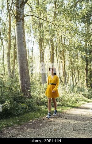 Giovane donna indossa marrone cappello, abito giallo e nero sneakers cercando e passeggiate nella foresta Foto Stock
