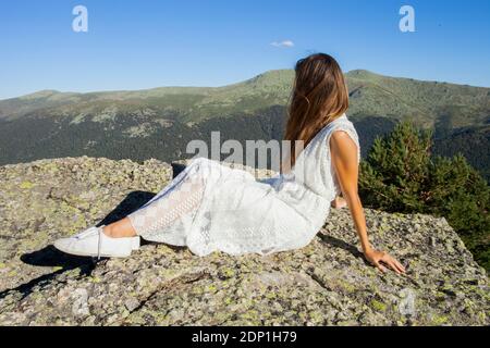 Donna bionda in abito bianco seduta su una grande roccia guarda la valle in una giornata di sole in estate Foto Stock