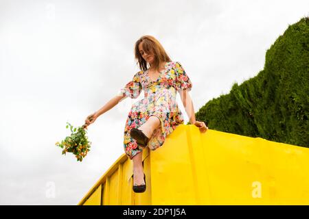 Donna in abito floreale seduta sul bordo del contenitore giallo, tenendo mazzo di fiori Foto Stock