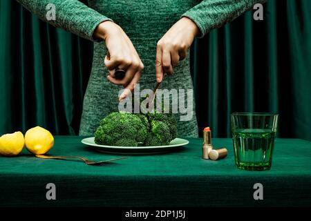 Metà sezione di donna che taglia broccoli con coltello e forchetta mentre in piedi contro la tenda verde Foto Stock