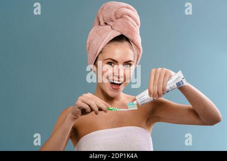 Bella ragazza eccitato che indossa asciugamani applicando dentifricio su spazzolino isolato su sfondo blu Foto Stock