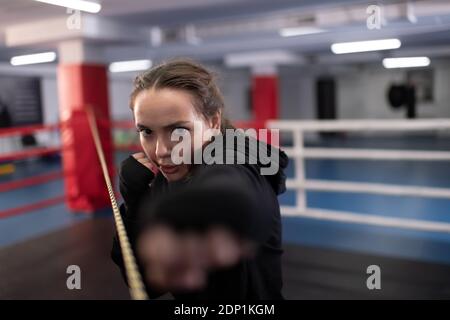 Serio combattente femmina facendo pugno jab e guardando la macchina fotografica mentre si pratica la boxe ombra durante l'allenamento in palestra contemporanea Foto Stock