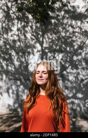 Giovane donna rossa in piedi con gli occhi chiusi contro l'ombra dell'albero parete Foto Stock