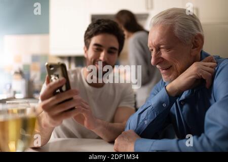 Anziano padre sorridente e guardando le foto su smartphone di felice giovane uomo seduto in cucina durante la cena di famiglia Foto Stock