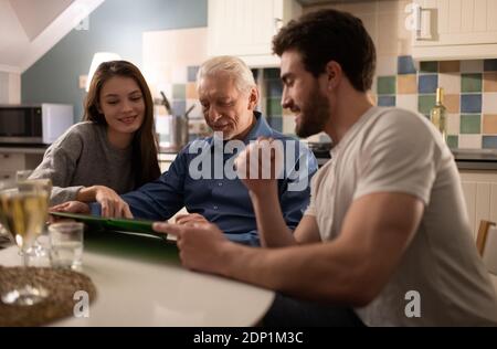 Giovane marito e moglie sorridono e guardano le foto in album di uomo anziano durante la cena di famiglia a casa Foto Stock