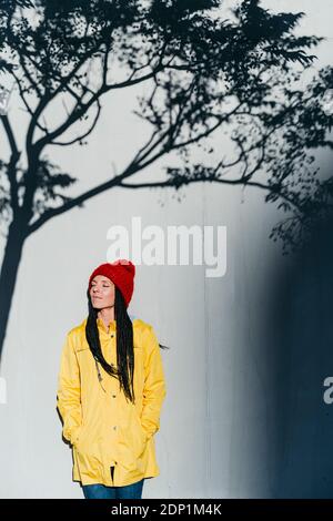Thoughtful woman wearing raincoat standing with hands in pockets under tree shadow against wall Stock Photo