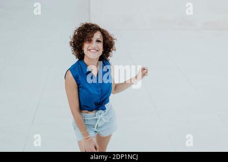 Donna sorridente con capelli ricci in piedi sul pavimento contro le piastrelle parete Foto Stock