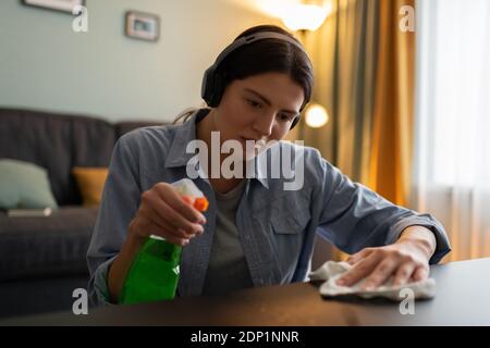Donna giovane e seria in camicia casual che ascolta musica con le cuffie e rimuovere le macchie dai mobili mentre si esegue la pulizia regolare a. casa Foto Stock