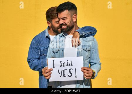 L'uomo gay abbracciando il giovane uomo che tiene carta con il testo i Ha detto Sì scritto su di esso contro parete gialla Foto Stock