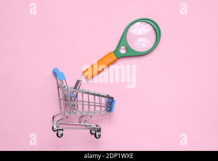 Lente di ingrandimento, carrello per lo shopping su sfondo rosa patel. Foto Stock