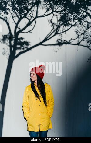 Donna che indossa un impermeabile in piedi con le mani in tasche sotto l'albero ombra contro il muro Foto Stock