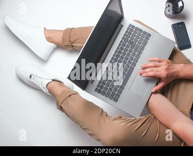 Frammento di gambe maschili in pantaloni beige e sneakers bianche seduta su bianco. Uomo che usa un computer portatile moderno. Lavoratore online, freelancing, lavoro a casa, lavoro uomo Foto Stock