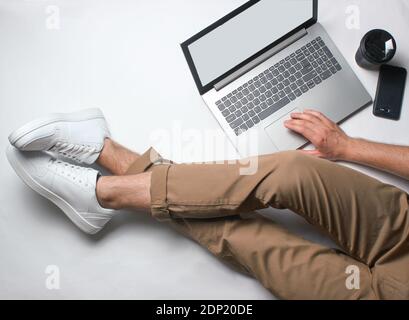 Frammento di gambe maschili in pantaloni beige e sneakers bianche seduta su bianco. Uomo che usa un computer portatile moderno. Lavoratore online, freelancing, lavoro a casa, lavoro uomo Foto Stock