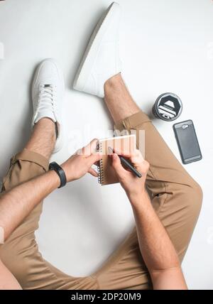 Lavoro di un giornalista. L'uomo scrive in un taccuino. Frammento di gambe maschili in pantaloni beige e sneakers bianche su sfondo bianco. Vista dall'alto. Foto Stock