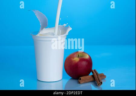 Una tazza generica di yogurt bianco, di plastica, su sfondo blu, con bastoncini di mela e cannella; spruzzi di yogurt Foto Stock