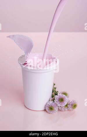 Una tazza generica, bianca, di plastica di yogurt rosa su sfondo rosa con i fiori rosa; spruzzi di yogurt Foto Stock