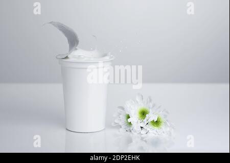 Una tazza generica, bianca, di plastica di yogurt bianco su sfondo bianco con i fiori bianchi; spruzzi di yogurt Foto Stock