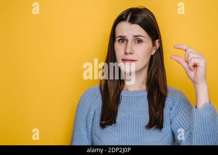 Bruna giovane donna isolato su sfondo giallo studio mostra molto minuscolo oggetto, forme qualcosa di piccolo, vestito in pullover blu Foto Stock